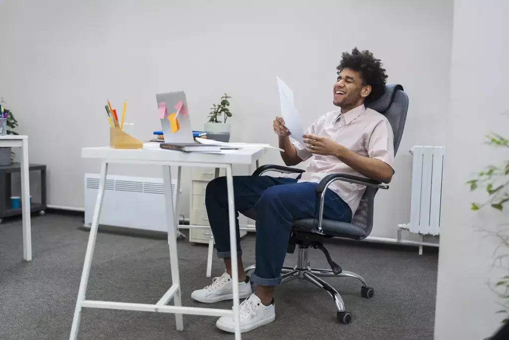  Young man sitting in an ergonomic home office chair at a desk, smiling and holding a piece of paper.